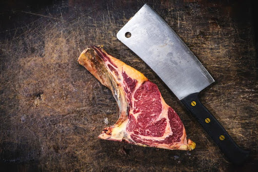 Raw tomahawk steak on a butcher block next to a large meat cleaver.