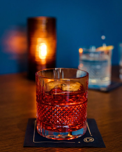 Red cocktail with an orange twist served in a textured glass on a coaster, with candlelight and other drinks in the background.