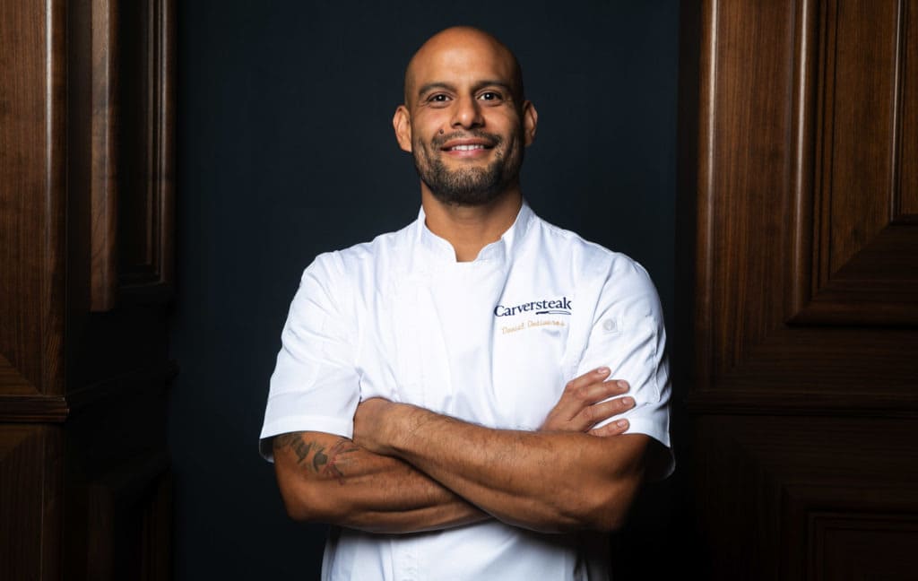 "Chef wearing a white Carversteak uniform standing confidently with arms crossed in front of a dark background and wood paneling."