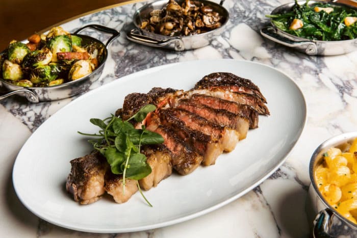 Sliced steak on a white plate with assorted vegetable sides on a marble table