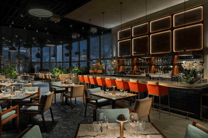 Upscale restaurant dining room with modern décor, orange bar stools, elegant table settings, and moody evening lighting.