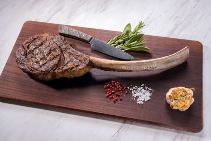 Tomahawk ribeye steak served on a dark wood cutting board, garnished with fresh herbs, roasted garlic, cracked salt, and peppercorns, accompanied by a sharp knife placed across the bone.