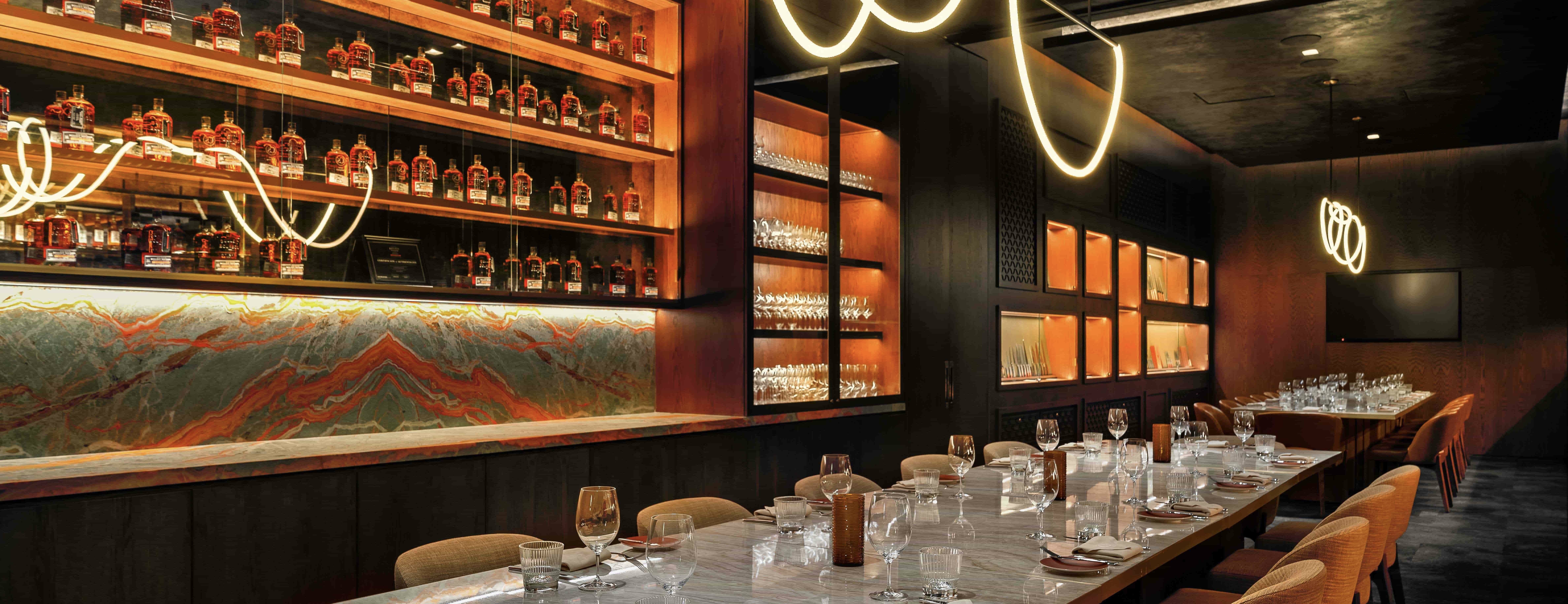 Elegant private dining room with a long marble table set for guests, surrounded by upholstered chairs, warm ambient lighting, and a wall display of liquor bottles and glassware.