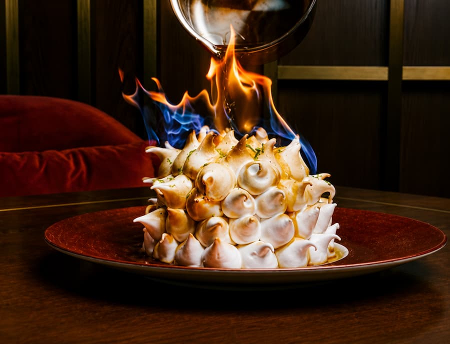 Flaming baked Alaska dessert topped with toasted meringue, served on a plate as a warm sauce is poured over the dessert, creating dramatic blue and orange flames.