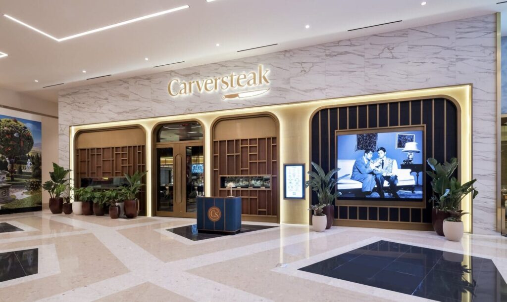 Carversteak entrance featuring illuminated signage, marble walls, decorative plants, and a digital display.