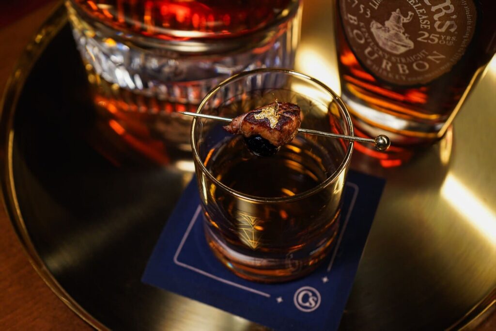 Bourbon cocktail garnished with a cherry and gold accent, served on a tray with whiskey bottles.