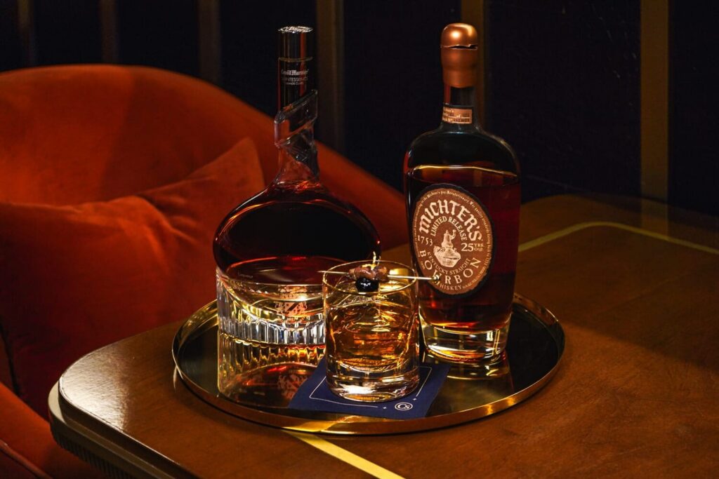 Whiskey cocktail displayed on a gold tray alongside two bottles of bourbon in a warmly lit setting.