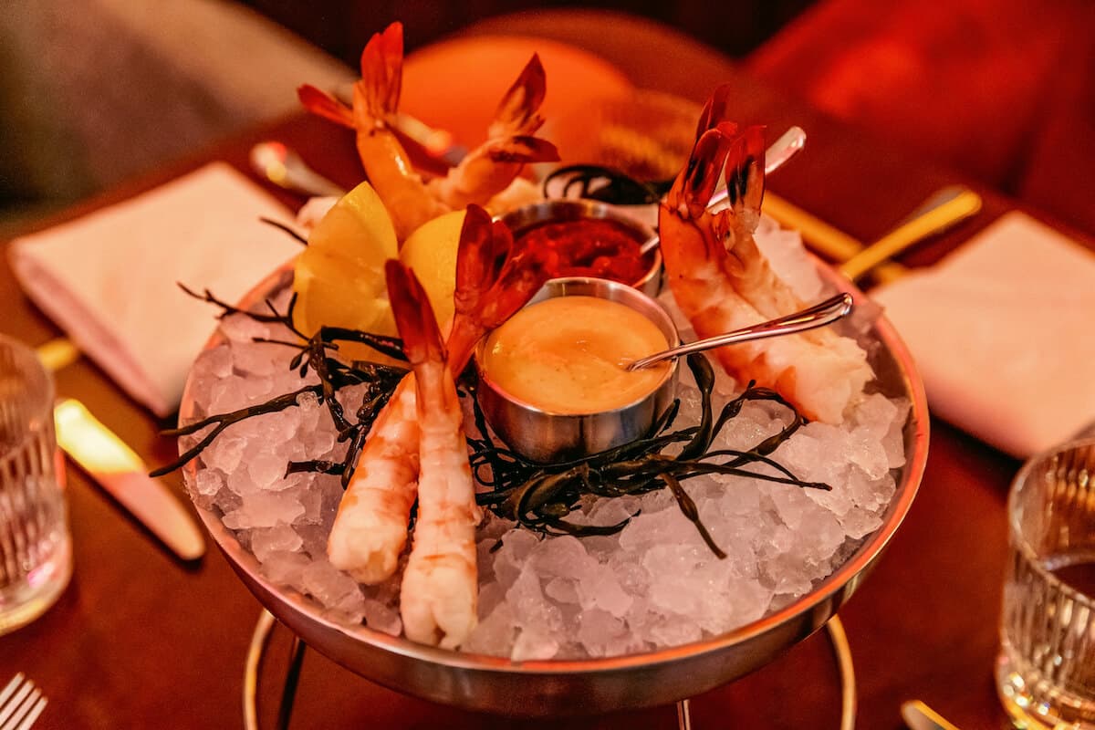 Chilled jumbo shrimp arranged on a bed of crushed ice in a metal serving bowl, accompanied by lemon wedges, cocktail sauce, and creamy dipping sauce under warm restaurant lighting.
